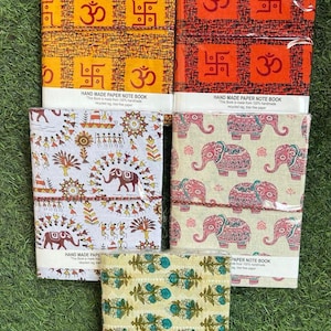 May include: A collection of handmade paper notebooks with various patterned covers. The designs include orange and yellow geometric patterns, elephant motifs, and floral prints. Each notebook is bound with a string.