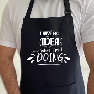 May include: Black apron with a large front pocket and adjustable neck strap. The apron features the white text, "I HAVE NO IDEA WHAT I'M DOING" in a whimsical font. The apron is worn by a person wearing a white t-shirt.