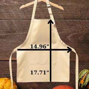 May include: A beige toddler apron, 14.96 inches wide and 17.71 inches long, hangs on a wooden hanger. The apron has a pocket and long ties. The word "Toddler" is written in red at the top. Gourds are at the bottom.