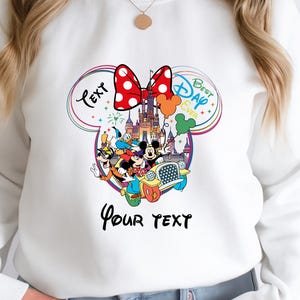 May include: White sweatshirt with a colorful graphic featuring Disney characters, a castle, and a red polka-dot bow. The graphic includes the text "Text" and "Best Day Ever" in a Mickey Mouse ear design, with space for custom text.