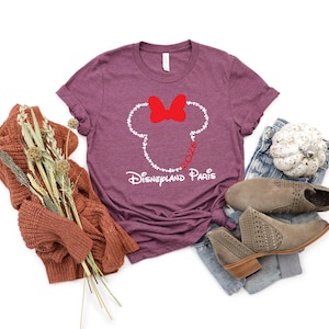 May include: A maroon t-shirt featuring a white outline of Minnie Mouse's head, a red bow, and the text "Disneyland Paris 2026." The shirt is paired with jeans, boots, and a fall-themed sweater.