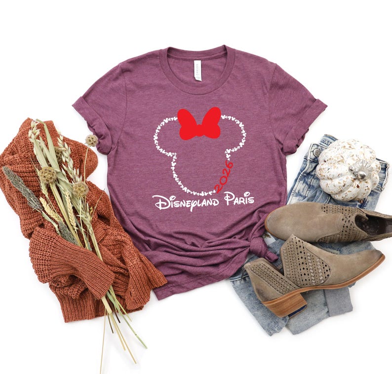 May include: A maroon t-shirt featuring a white outline of Minnie Mouse's head, a red bow, and the text "Disneyland Paris 2026." The shirt is paired with jeans, boots, and a fall-themed sweater.