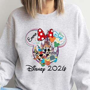 May include: A light gray sweatshirt featuring a colorful Disney-themed graphic. The design includes Mickey and Minnie Mouse, Donald Duck, and Goofy in a car, with a castle, balloons, and the text "Disney 2020". A red bow with white polka dots is at the top.