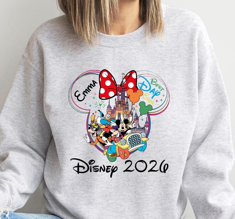 May include: A light gray sweatshirt featuring a colorful Disney-themed graphic. The design includes Mickey and Minnie Mouse, Donald Duck, and Goofy in a car, with a castle, balloons, and the text "Disney 2020". A red bow with white polka dots is at the top.
