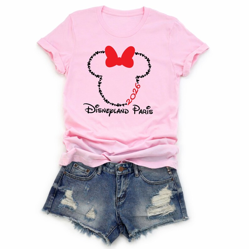 May include: A light pink t-shirt with a graphic of a Minnie Mouse head made of black hearts and a red bow. The text "Disneyland Paris" and "2026" are also printed on the shirt. The shirt is paired with denim shorts.
