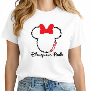 May include: White t-shirt featuring a Minnie Mouse design. The design includes a red bow, a silhouette of Minnie's head made of black dots, and the text "Disneyland Paris 2026" in black and red.