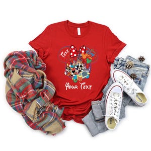 May include: A red t-shirt featuring a Disney-themed graphic with Mickey and Minnie Mouse, a castle, and the text "Best Day Ever" and "Your Text". The shirt is paired with blue jeans, white sneakers, and a plaid scarf.