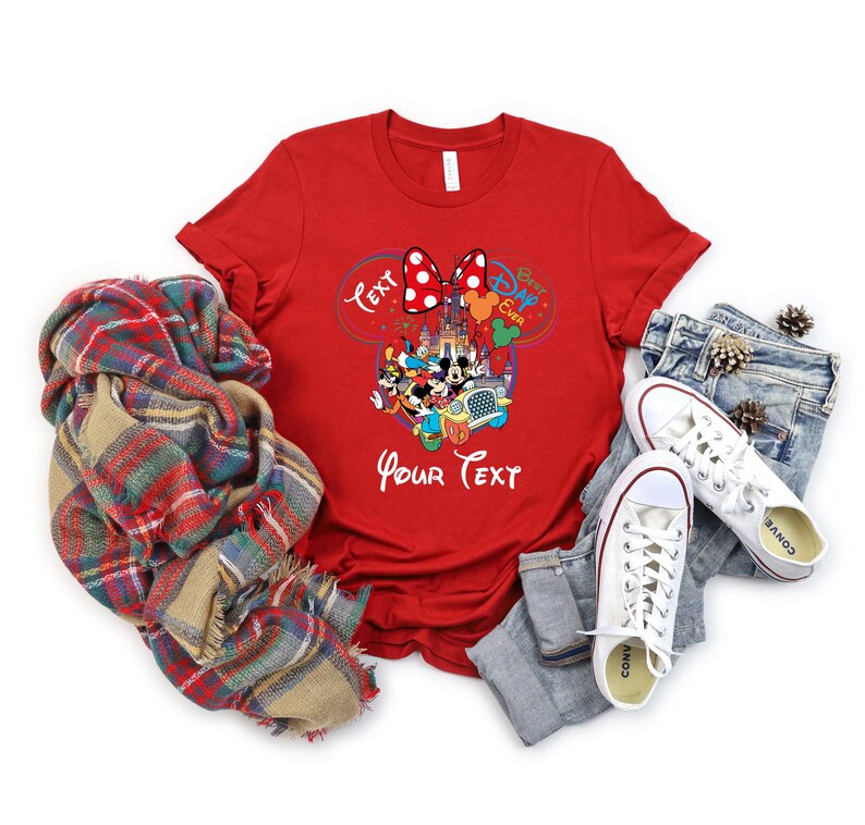 May include: A red t-shirt featuring a Disney-themed graphic with Mickey and Minnie Mouse, a castle, and the text "Best Day Ever" and "Your Text". The shirt is paired with blue jeans, white sneakers, and a plaid scarf.