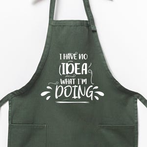 May include: A dark green apron with two front pockets and long ties. The apron features the white text, "I HAVE NO IDEA WHAT I'M DOING" in a whimsical font. The apron is hanging against a white background.