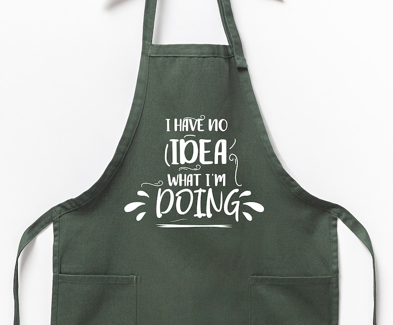 May include: A dark green apron with two front pockets and long ties. The apron features the white text, "I HAVE NO IDEA WHAT I'M DOING" in a whimsical font. The apron is hanging against a white background.