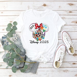 May include: White t-shirt featuring a colorful Disney graphic with Mickey and Minnie Mouse, a castle, and the text "Disney 2025." The design includes the name "Olivia" and the words "Best Day Ever." The shirt is paired with jeans and white sneakers.