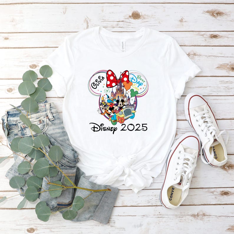 May include: White t-shirt featuring a colorful Disney graphic with Mickey and Minnie Mouse, a castle, and the text "Disney 2025." The design includes the name "Olivia" and the words "Best Day Ever." The shirt is paired with jeans and white sneakers.