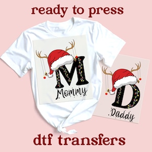 Könnte beinhalten: Weißes T-Shirt mit einem weihnachtlichen Design. Das Design zeigt den Buchstaben "M" mit Weihnachtsmütze, Geweih und dem Wort "Mommy". Ein separates Design zeigt den Buchstaben "D" mit Weihnachtsmütze, Geweih und dem Wort "Daddy". Der Text "ready to press" und "dtf transfers" sind ebenfalls sichtbar.