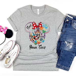 May include: A gray t-shirt with a Disney-themed graphic, featuring Mickey and Minnie Mouse, a castle, and the text "Best Dad" and "Your Text". The image also includes a Minnie Mouse ear headband, white sneakers, and blue jeans.