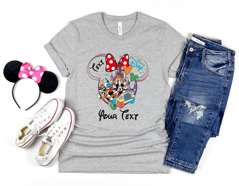 May include: A gray t-shirt with a Disney-themed graphic, featuring Mickey and Minnie Mouse, a castle, and the text "Best Dad" and "Your Text". The image also includes a Minnie Mouse ear headband, white sneakers, and blue jeans.