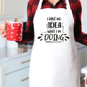 May include: A white apron with the text "I HAVE NO IDEA WHAT I'M DOING" in black script. A red mug with white heart shapes is held in the left hand. The person is wearing a red and black plaid shirt.