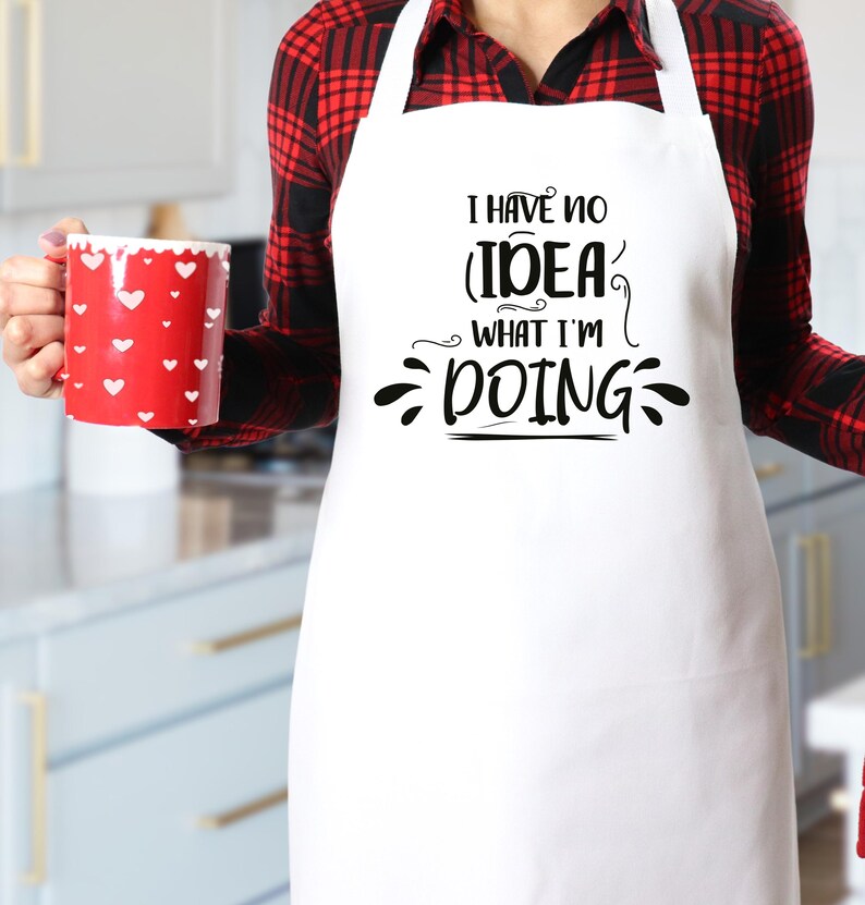 May include: A white apron with the text "I HAVE NO IDEA WHAT I'M DOING" in black script. A red mug with white heart shapes is held in the left hand. The person is wearing a red and black plaid shirt.