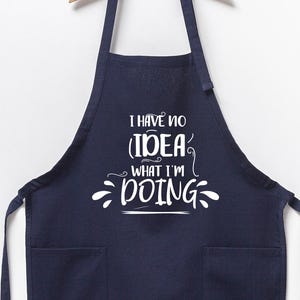 May include: Navy blue apron with two front pockets and the text "I HAVE NO IDEA WHAT I'M DOING" in white script. The apron is hanging from a wooden hanger against a white background. A kitchen accessory.