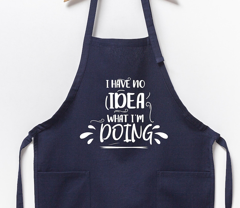 May include: Navy blue apron with two front pockets and the text "I HAVE NO IDEA WHAT I'M DOING" in white script. The apron is hanging from a wooden hanger against a white background. A kitchen accessory.