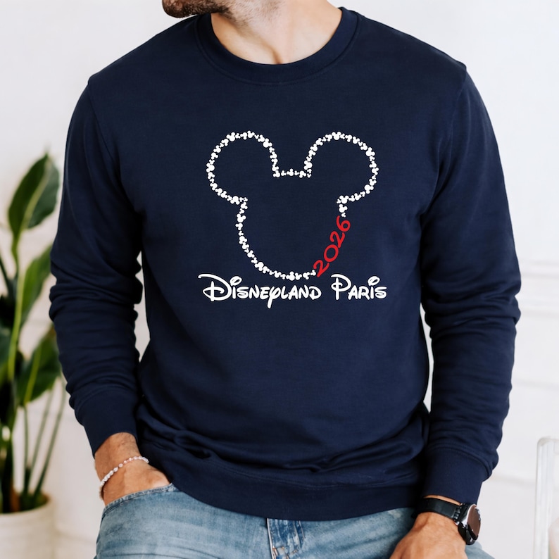 May include: Navy blue sweatshirt featuring a white Mickey Mouse head outline made of small hearts. The text "2026" in red and "Disneyland Paris" in white are below the design. The sweatshirt is a long-sleeved crew neck.
