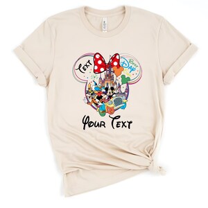 May include: Cream-colored t-shirt featuring a colorful Disney graphic. The design includes Mickey and Minnie Mouse, a castle, and the text "Best Day Ever" and "Your Text". A red polka-dot bow is at the top of the design.