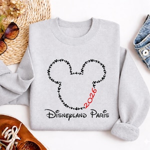 May include: A light gray sweatshirt featuring a Mickey Mouse head outline made of small black hearts. The number "2026" is in red, and the words "Disneyland Paris" are printed in black below the design. The sweatshirt is made of a soft material.
