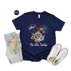 May include: Navy blue t-shirt featuring a colorful graphic of Disney characters in front of a castle. The shirt includes the text "Mike" and "Best Dad Ever" inside Mickey Mouse ears, and "The Hill Family" below the graphic. A pair of white sneakers and denim shorts are next to the shirt.