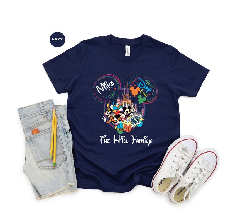 May include: Navy blue t-shirt featuring a colorful graphic of Disney characters in front of a castle. The shirt includes the text "Mike" and "Best Dad Ever" inside Mickey Mouse ears, and "The Hill Family" below the graphic. A pair of white sneakers and denim shorts are next to the shirt.