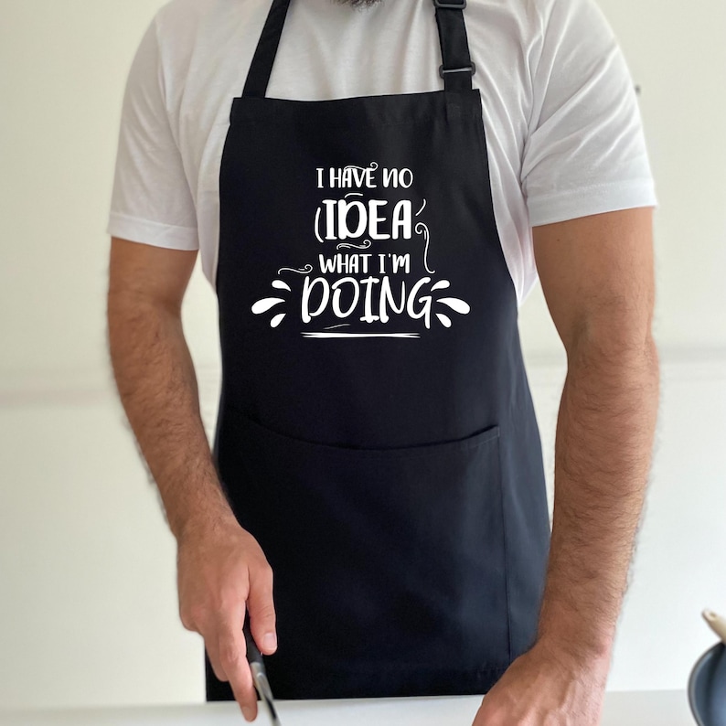 May include: Black apron with a large front pocket and adjustable neck strap. The apron features the white text, "I HAVE NO IDEA WHAT I'M DOING" in a whimsical font. The apron is worn by a person wearing a white t-shirt.
