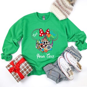 May include: Green sweatshirt featuring a Minnie Mouse bow and a Disney character design. The design includes the text "Text" and "Your Text". A gift box with a red bow and a winter hat are also in the image.
