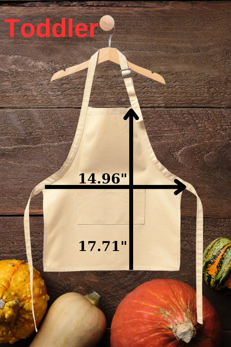 May include: A beige toddler apron, 14.96 inches wide and 17.71 inches long, hangs on a wooden hanger. The apron has a pocket and long ties. The word "Toddler" is written in red at the top. Gourds are at the bottom.