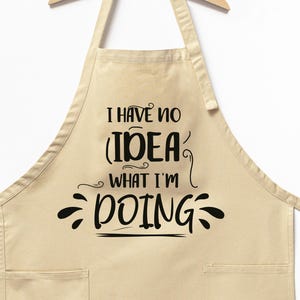 May include: A beige apron with two front pockets and the text "I HAVE NO IDEA WHAT I'M DOING" in black script. The apron is hanging on a wooden hanger against a white background.