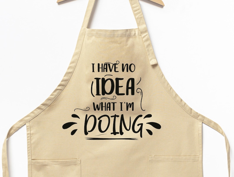 May include: A beige apron with two front pockets and the text "I HAVE NO IDEA WHAT I'M DOING" in black script. The apron is hanging on a wooden hanger against a white background.