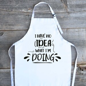 May include: White apron with the text "I HAVE NO IDEA WHAT I'M DOING" in black script. The apron has a neck strap and waist ties. The apron is displayed on a wooden surface.
