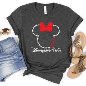 May include: Dark gray t-shirt featuring a Minnie Mouse design with a red bow and the text "Disneyland Paris 2026". The shirt is paired with denim shorts, sunglasses, and gold sandals. A casual, travel-themed outfit.