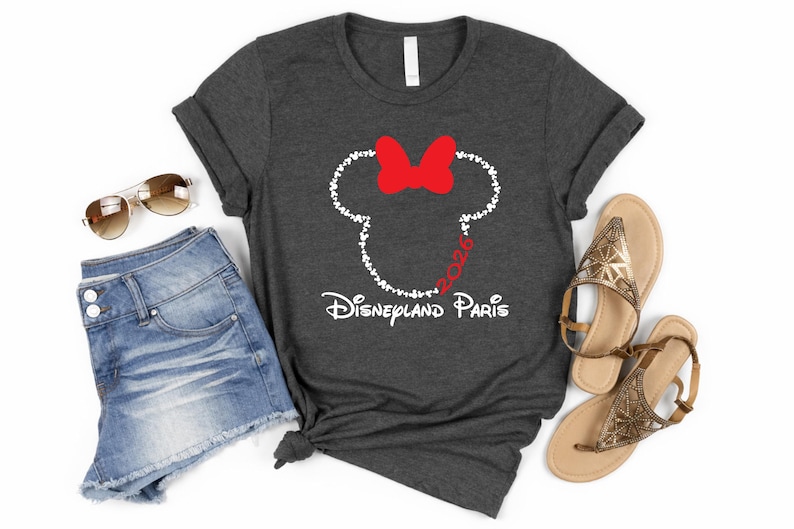 May include: Dark gray t-shirt featuring a Minnie Mouse design with a red bow and the text "Disneyland Paris 2026". The shirt is paired with denim shorts, sunglasses, and gold sandals. A casual, travel-themed outfit.