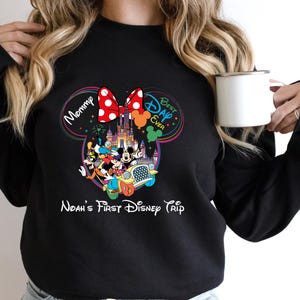 May include: Black sweatshirt featuring a colorful Disney-themed graphic. The design includes Mickey and Minnie Mouse, a castle, and the text "Mommy," "Best Day Ever," and "Noah's First Disney Trip." A white mug is held in the hand.
