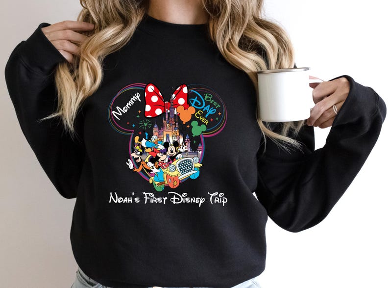 May include: Black sweatshirt featuring a colorful Disney-themed graphic. The design includes Mickey and Minnie Mouse, a castle, and the text "Mommy," "Best Day Ever," and "Noah's First Disney Trip." A white mug is held in the hand.