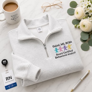 May include: A light gray quarter-zip sweatshirt featuring embroidered text that reads Gaizel, MS, BCBA and Board Certified Behavioral Analyst, accompanied by a row of four small, colorful stick figures in blue, pink, purple, and yellow.