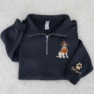 May include: A black quarter-zip sweatshirt featuring an embroidered brown and white dog wearing a purple witch hat and holding a jack-o'-lantern. The sleeve displays an embroidered tan paw print above the name Burger in tan cursive script.