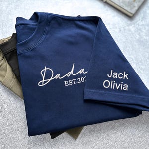 May include: A navy blue t-shirt featuring white embroidered text that reads Dada EST.2024 on the chest and the names Jack and Olivia on the sleeve. The shirt is folded and layered over other shirts in olive green and black colors on a textured surface.