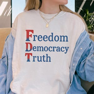 Könnte beinhalten: Ein cremefarbenes T-Shirt mit den Worten "Freedom Democracy Truth" in roter und blauer Schrift. Das Shirt wird mit einer hellblauen Jeansjacke und einer blauen Jeans kombiniert. Eine schwarze Handtasche ist sichtbar.