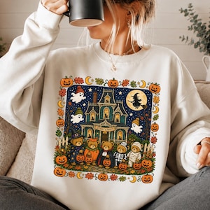 Pode incluir: Uma camisola de gola redonda cor de creme com uma impressão gráfica colorida de uma casa assombrada, fantasmas e cinco ursos de peluche vestidos com fatos de Halloween. O texto "SPOOKY & SWEET" está impresso na casa. O design é emoldurado por abóboras.