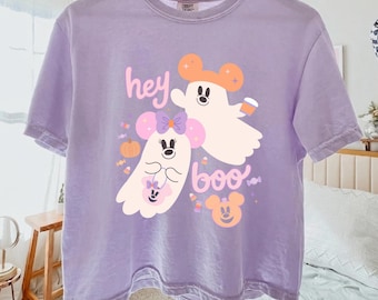 May include: A light purple t-shirt featuring a graphic of two white ghosts with mouse ears, one wearing a purple bow and holding a small bag, the other holding a coffee cup. The text "hey boo" is printed in pink and white with candy icons.