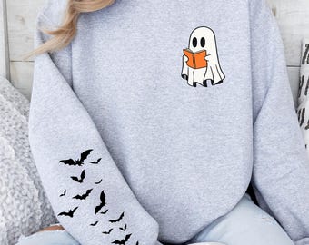 May include: A light gray crewneck sweatshirt featuring a small graphic of a white ghost reading an orange book on the left chest and a cluster of black bats printed along the lower portion of the right sleeve, made of a soft cotton blend fabric.