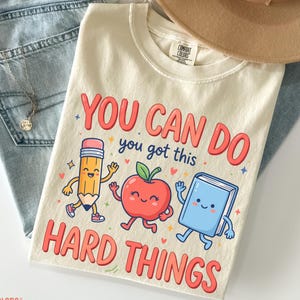 May include: An ivory t-shirt featuring a graphic with a cartoon pencil, apple, and book. The text reads, "YOU CAN DO you got this HARD THINGS." The shirt is displayed with light blue denim jeans, a tan wide-brimmed hat, and a silver necklace.