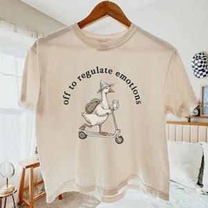 Puede incluir: Una camiseta de manga corta color crema con el gráfico de un ganso que lleva un sombrero y una mochila mientras monta en un patinete y sostiene una taza de café. El texto "off to regulate emotions" aparece en un arco oscuro sobre el ganso.