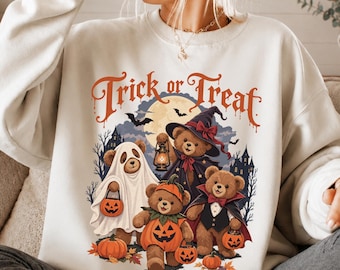 May include: A cream-colored crewneck sweatshirt featuring a graphic print with the text Trick or Treat. The design displays four teddy bears in Halloween costumes, including a ghost, a witch, a pumpkin, and a vampire, surrounded by pumpkins.