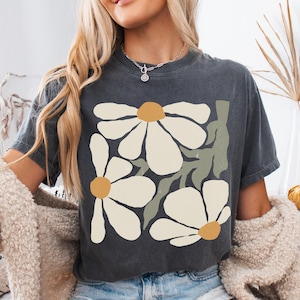 Puede incluir: Camiseta gris oscuro con un dise&ntilde;o floral de grandes margaritas blancas, centros amarillos y tallos verdes. La camiseta est&aacute; hecha de material suave y es adecuada para ropa casual. La camiseta se combina con pantalones cortos de mezclilla.