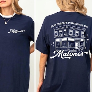 Op de afbeelding: Marineblauw T-shirt met witte bies en het woord "Malone's" in een sierlijk lettertype op de voorkant. De achterkant toont een witte illustratie van een restaurant met de tekst "Best Burgers in Hastings, MA!" en "Malone's".
